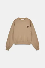 Sudadera de algodón color beige, con cuello redondo, manga larga y corte ligeramente corto. Presenta un pequeño parche cuadrado marrón con un logo grabado en el pecho.