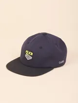Gorra de lona tipo snapback color azul marino con visera plana negra. Presenta un parche frontal con diseño gráfico en tonos amarillo y blanco, y el logo de la marca bordado en el lateral.
