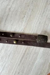 Cinturón de gamuza color chocolate con hebilla metálica rectangular de estilo vintage, decorada con relieve floral y pequeñas incrustaciones de piedras turquesas. Presenta detalles de tachas metálicas circulares a lo largo de la tira.