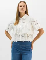 Camisa de algodón blanca con cuello clásico y cierre frontal de botones. Presenta un diseño con capas superpuestas y volados, combinando paneles de tela lisa con tejido bordado suizo de motivos calados y una cenefa decorativa. Posee mangas cortas y una silueta acampanada.