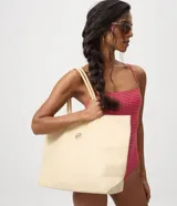 Cartera amplia confeccionada en fibra de papel, de color beige, con asas de hombro, cierre superior y detalle metálico dorado claro en el frente.
