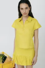 Top de lino color amarillo, con cuello camisero, cierre frontal parcial y mangas cortas. Presenta pespuntes decorativos a contratono.