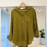 Camisa verde pistacho de manga larga, con cuello solapa, cierre frontal de botones, bolsillo en el pecho y diseño de corte asimétrico más largo en la parte trasera.
