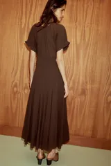 Vestido midi de color marrón, confeccionado en tejido de gasa con textura. Presenta un escote en pico con detalle de lazada frontal, mangas cortas abullonadas con terminación elástica y falda con detalle de godets que aportan volumen. Incluye forro interior a tono y cierre lateral con cremallera oculta.