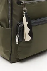 Mochila urbana de nylon en color verde militar, con detalles en cuero sintético negro. Cuenta con compartimento principal con cierre, bolsillo frontal con cierre, bolsillos laterales abiertos y asa superior. Incluye un colgante decorativo de tela y correas ajustables acolchadas.