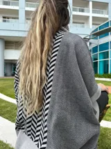 Poncho oversize con cuello alto negro, diseño geométrico en blanco y negro, bolsillo lateral y detalle de bloques de color en gris y negro.