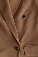 Blazer color marrón levemente largo con solapas de muesca, hombreras y doble botonadura frontal. Cuenta con bolsillos delanteros ribeteados con solapa y diseño forrado.