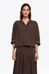 Camisa corta de corte boxy en color marrón, con cuello camisero, cierre frontal de botones y mangas 3/4. Presenta pespuntes decorativos a contraste en los puños y una textura sutil en el tejido.