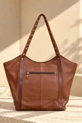 Cartera tipo tote de gamuza color azul marino, con asas dobles de cuero marrón texturado y detalles de tachas metálicas plateadas. Cuenta con una placa metálica frontal con el logo de la marca Pecarí.