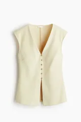 Chaleco de punto interlock color crema, entallado, con escote en V pronunciado y manga japonesa. Presenta una hilera de botones metálicos pequeños en el frente que generan un ligero corte acampanado en la parte inferior.