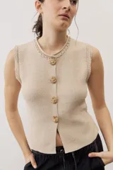 Chaleco de punto color beige sin mangas, con cuello redondo y cierre frontal de botones dorados con diseño texturizado en forma de sol.