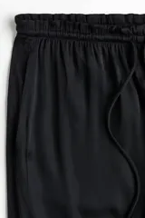 Pantalón holgado de satén negro, con piernas amplias, cintura elástica con cordón ajustable y bolsillos laterales.