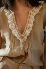 Camisa de lino color beige con escote en V profundo, mangas con volados y detalles de puntilla color crudo en el escote y los hombros. Presenta cierre frontal con botones forrados y un corte relajado.