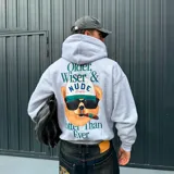 Hoodie gris de corte oversize con capucha y bolsillo frontal tipo canguro. Presenta un estampado gráfico en la espalda de un oso con anteojos de sol, gorra y un cigarro, acompañado por la frase 'Older, Wiser & Nude Project Hotter Than Ever'.