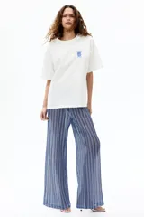 Remera oversized de algodón blanco, con cuello redondo acanalado, mangas cortas con hombros caídos y pequeño estampado azul en el pecho de un jarrón con flores y el número 1096.