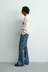 Sudadera de tejido con algodón, color beige claro, de corte relajado (relaxed fit) y cuello redondo. Presenta un parche bordado en el pecho con el logo de Ford y el texto "Genuine Parts" en azul marino.