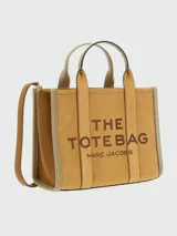 Cartera tipo tote de Marc Jacobs, confeccionada en gamuza color tostado con detalles en cuero marrón. Presenta el logo característico estampado en el frente, doble asa superior y correa ajustable extraíble.