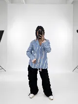 Camisa de corte oversize con estampado de rayas verticales en tonos azul y blanco. Presenta un diseño distintivo de doble puño y cuello en color rosado contrastante, cierre frontal con botones y un pequeño detalle de etiqueta en el frente.