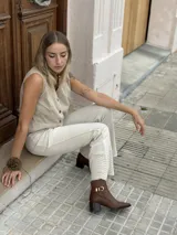 Bota corta de cuero color marrón, con taco cuadrado de madera y punta fina. Presenta una tira decorativa con hebilla metálica dorada alrededor del tobillo.
