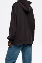 Buzo con capucha de algodón y poliéster, color negro lavado (acid wash), con bolsillo canguro y cordones rojos. Presenta un estampado frontal en plateado con un lazo y el texto "BIMBA Y LOLA".