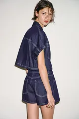 Camisa corta de denim de manga corta, con cuello solapa, escote en V y bolsillos delanteros de plastrón. Cuenta con costuras en contraste y cierre frontal con botones metálicos.