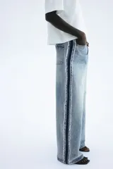 Jean de tiro alto, confeccionado en denim de algodón rígido, de corte ancho y pierna recta. Presenta un lavado celeste claro con efecto desgastado en el centro y franjas laterales en denim más oscuro con acabado desflecado.