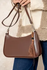 Cartera tipo baguette de diseño minimalista, confeccionada en una combinación de gamuza color beige en el panel frontal y cuero color chocolate en los bordes y la correa. Incluye un detalle colgante de cuentas de madera y cierre superior.