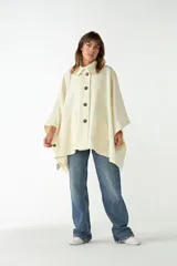 Capa de lana fina sin teñir, color crema, con cuello camisero, cierre frontal de botones oscuros y dos bolsillos grandes aplicados en la parte delantera.