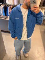 Campera canguro de algodón azul con cierre frontal, capucha y bolsillos tipo parche. Presenta un pequeño bordado ovalado en el pecho.