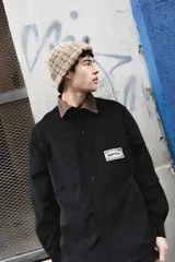 Sobrecamisa negra de corte clásico con cuello camisero en contraste color marrón a cuadros. Presenta cierre frontal con botones, dos bolsillos frontales tipo parche y una etiqueta decorativa con el logo de la marca en el pecho.