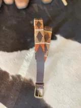 Cinturón de cuero en tono tostado con bordado a mano de hilos encerados en tonos marrones y beige con motivos geométricos, y hebilla metálica rectangular.