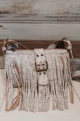 Mini bandolera de cuero con diseño de flecos largos y hebilla frontal decorativa. Presenta un acabado texturizado con efecto craquelado en tonos blanco y marrón, y correa ajustable.