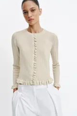Chaqueta de punto canalé en color beige con hilo metalizado, cuello redondo, manga larga y cierre frontal con botones. Presenta detalles de volantes en la tapeta de los botones, el ruedo y los puños.