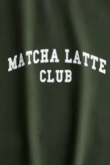 Remera de manga raglán con cuerpo en color verde oscuro y mangas en color blanco. Presenta cuello redondo y estampado frontal con la inscripción 'MATCHA LATTE CLUB' en tipografía universitaria.