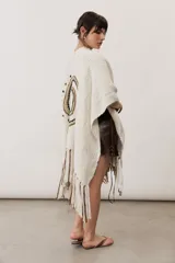 Ruana de punto color beige con detalles geométricos negros y flecos largos tricolores en el borde inferior.