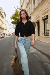 Chaqueta corta negra de tejido texturizado, con cuello redondo y manga ancha tipo kimono que termina por debajo del codo. Cierre frontal con un único botón joya metálico plateado con relieve.