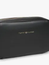 Cartera tipo bandolera Tommy Hilfiger de color negro, con acabado liso, diseño compacto y esquinas redondeadas. Cuenta con cierre de cremallera superior, inscripción y logotipo de la marca en bajo relieve dorado en el frente, y una correa cruzada ancha ajustable de jacquard a rayas en blanco y negro con la marca escrita.