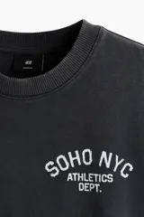Remera boxy de algodón gris oscuro, con corte oversized, hombros caídos, cuello redondo ribeteado y estampa pequeña en el pecho con texto en blanco.