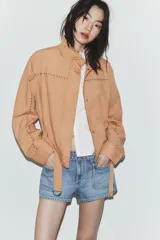 Chaqueta color tostado con cuello subido, manga larga y detalle de tachas metálicas en contraste. Incluye cinturón del mismo tejido con hebilla metálica y cierre frontal con cremallera y botones a presión ocultos.