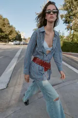 Vestido corto de manga larga con cuello solapa y cierre frontal de botones a presión. Presenta un diseño a cuadros en tonos azul y marrón, cintura ajustable con cinturón de hebilla y falda con volantes en el bajo.