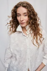 Vestido camisero corto y de silueta amplia en color blanco, con cuello clásico, cierre frontal de botones y mangas largas. Cuenta con diseño bordado floral en toda la prenda y dobladillo asimétrico, más largo en la parte trasera.