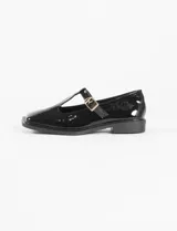 Mocasín estilo T-bar color negro en charol, con punta cuadrada, tira en el empeine con hebilla dorada y suela plana.