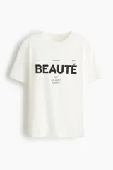 Remera de algodón blanca con cuello redondo, mangas cortas y estampa frontal con la frase "À la recherche de la beauté. Un regard suffit" en tipografía negra.