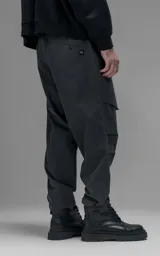 Pantalón de estilo utilitario confeccionado en textil técnico ripstop color gris oscuro. Presenta un corte oversize con cintura elástica, cierre frontal con broche técnico, bolsillos delanteros de gran tamaño con cierre y sistema de ajuste en los tobillos mediante broches snap.