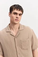Camisa de manga corta color beige confeccionada en muselina de algodón, con cuello abierto, cierre frontal de botones y un bolsillo aplicado en el pecho.