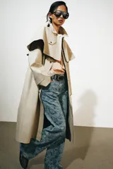 Gabardina larga de corte recto y diseño minimalista en color beige, con cuello solapa, manga larga y cierre frontal con botones. Cuenta con una capa desmontable en el mismo tejido y una solapa decorativa en el pecho.