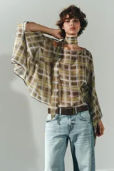 Capa corta con estampado a cuadros en tonos mostaza, marrón y blanco. Presenta un cuello redondo y mangas amplias tipo murciélago. Incluye un lazo o foulard integrado en el cuello para anudar.