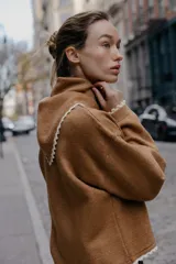 Jersey de punto color marrón, manga larga y corte holgado, con cuello tipo bandana anudado y terminaciones con puntilla blanca a contraste.