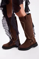 Bota de caña alta confeccionada en cuero vacuno, color negro, con diseño de tachas metálicas dispuestas en forma de arco a lo largo de la caña y correa decorativa con hebilla en la zona del tobillo. Presenta suela track de goma robusta.