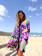 Camisero corto de corte holgado con estampado floral vibrante en tonos lila, fucsia, verde y negro. Presenta cierre frontal con botones, cuello camisero y mangas cortas.